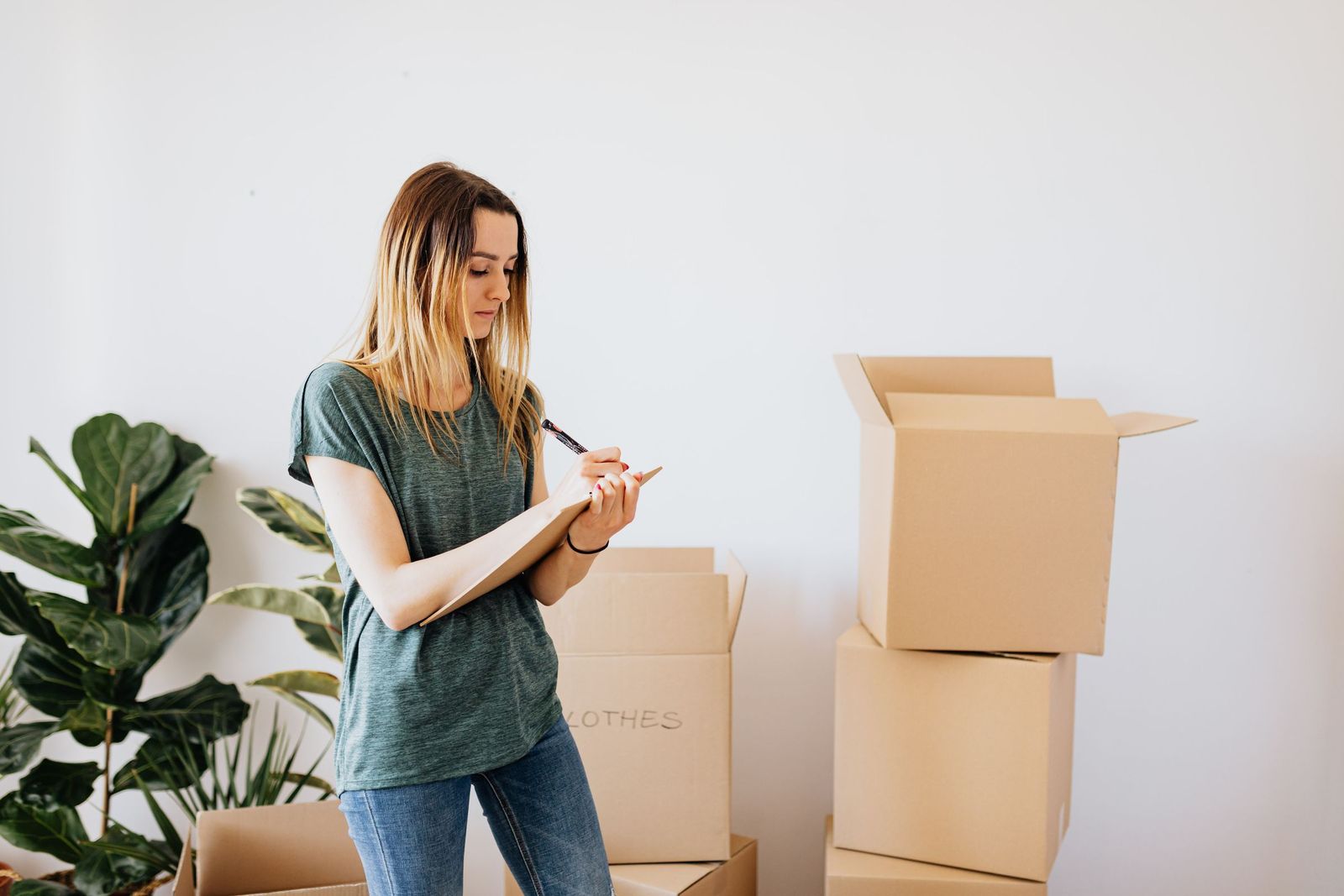 A woman writes notes on a clipboard beside packed cardboard boxes, listing what her house move will cost.