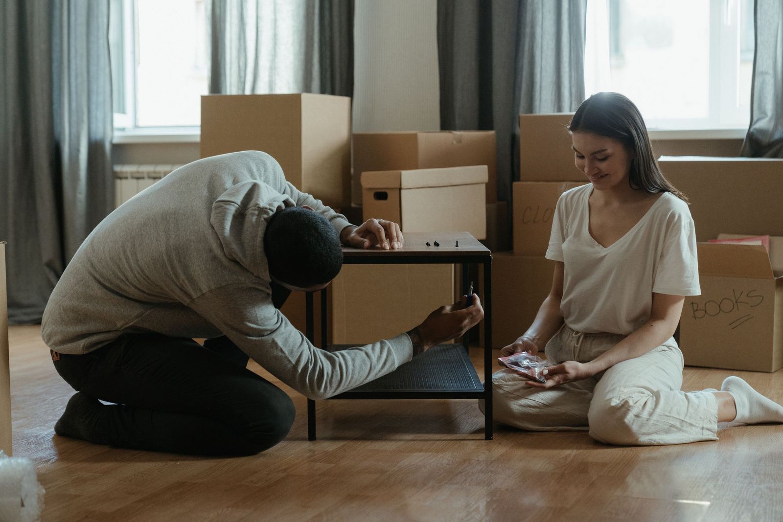 A couple kneeling on a wooden floor assembling a flat-pack side table, surrounded by stacked cardboard boxes in their new home.